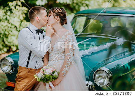 Just married wedding couple is standing near the retro vintage car in the park. Summer sunny day in forest. bride in elegant white dress with bouquet and elegant groom in love hugging. Bride and groom Just married wedding couple is standing near the retro vintage car in the park. Summer sunny day in forest. bride in elegant white dress with bouquet and elegant groom in love hugging. Bride and groom 85271806