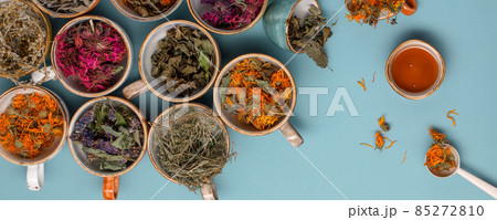 Assortment of dried relaxing tea herbs in colourful cups on blue background with honey close up. Calendula, mint, anise hyssop, monarda didyma, wormwood, sage leaves. Assortment of dried relaxing tea herbs in colourful cups on blue background with honey close up. Calendula, mint, anise hyssop, monarda didyma, wormwood, sage leaves. 85272810