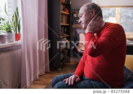 Frustrated senior man in red sweater holding hand on his neck sitting on sofa Frustrated senior man in red sweater holding hand on his neck sitting on sofa 85275098