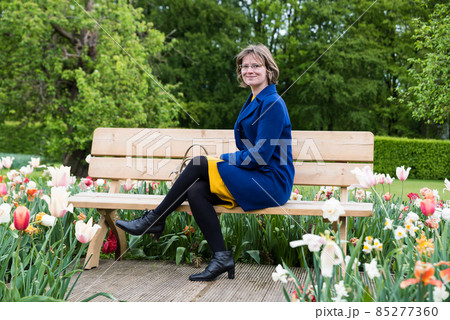 Attractive 30 year old woman sitting on a wooden bench, surrounded by blooming flowers 85277360