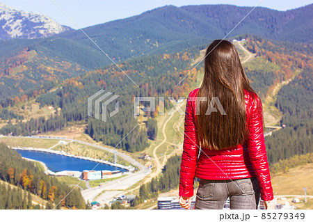 Beautiful cute slender girl in red down jacket, gray jeans staying on a top of a hill enjoying nature. Mountains, pond or river view in autumn sunny day. Young woman traveler back view, faceless trend 85279349