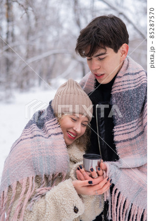 A young couple walks in the park in winter. The guy and the girl are drinking a warming drink outdoors. 85287570