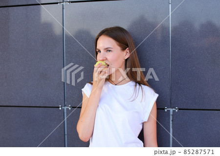 Young beautiful girl holds in hand and bites small green apple against background of gray wall. Young beautiful girl holds in hand and bites small green apple against background of gray wall. 85287571