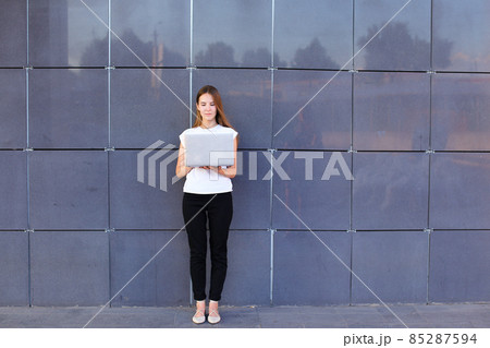 Perspective young girl entrepreneur holds laptop and works, solves problems, and stands on background wall of business center. Perspective young girl entrepreneur holds laptop and works, solves problems, and stands on background wall of business center. 85287594