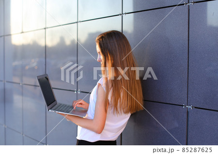 Attractive successful woman entrepreneur keeps in hands computer and works, typesets text, resolves issues on background of wall of business center. 85287625