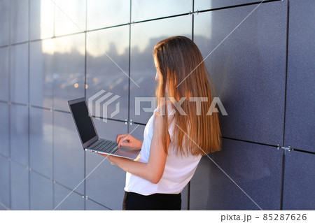 Attractive successful woman entrepreneur keeps in hands computer and works, typesets text, resolves issues on background of wall of business center. 85287626