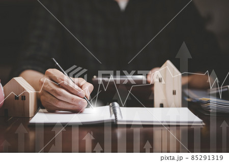 Businessman hand write in book note and icon business graphs with mini wood house model from model on wood table, Planning buy Real Estate, Planning to buy property. concept of Choose the best. 85291319