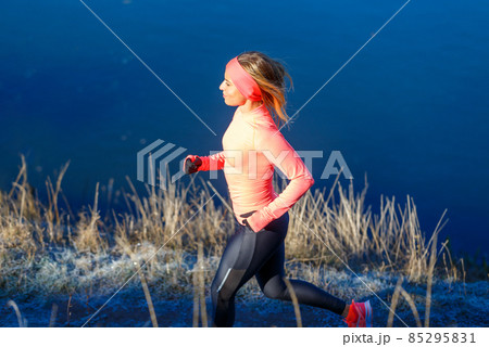 Young sporty woman in orange longsleeve running along blue frosty lake in the morning Young sporty woman in orange longsleeve running along blue frosty lake in the morning 85295831