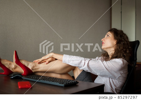 Tired young woman with curly hair and white shirt is working at the office with her computer, sitting with legs in red high heels at the table, routine work, freelance, burnout syndrome Tired young woman with curly hair and white shirt is working at the office with her computer, sitting with legs in red high heels at the table, routine work, freelance, burnout syndrome 85297297