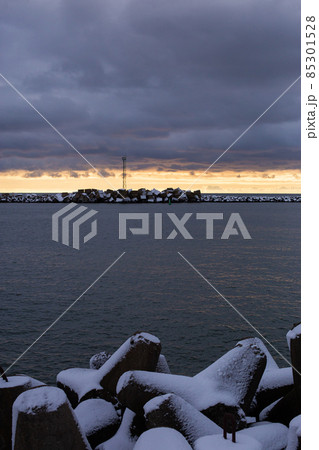 Evening on Curonian spit pier with protecting barrier rocks. Heavy dramatic sky, dark clouds and a line of bright yellow sunset light. Vertical photo Evening on Curonian spit pier with protecting barrier rocks. Heavy dramatic sky, dark clouds and a line of bright yellow sunset light. Vertical photo 85301528