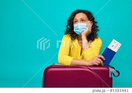 Beautiful upset woman in yellow cloth, wearing protective medical mask, holds passport with ticket and boarding pass, leans on her luggage and looks dreamily to the side. Blue background copy ad space Beautiful upset woman in yellow cloth, wearing protective medical mask, holds passport with ticket and boarding pass, leans on her luggage and looks dreamily to the side. Blue background copy ad space 85303461