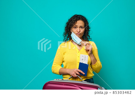 Sad young woman in bright yellow sweater takes off her protective medical mask and sadly looks at camera, posing against blue background, holding suitcase, air ticket and boarding pass with passport 85303462