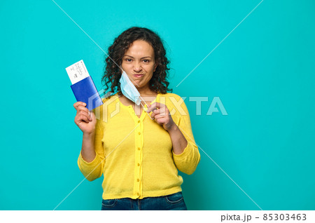 Upset and sad young woman traveler in yellow sweater puts her medical mask off and grimaces looking at camera, holding a boarding pass, air flight ticket and passport. Copy ad space. Travel concept Upset and sad young woman traveler in yellow sweater puts her medical mask off and grimaces looking at camera, holding a boarding pass, air flight ticket and passport. Copy ad space. Travel concept 85303463