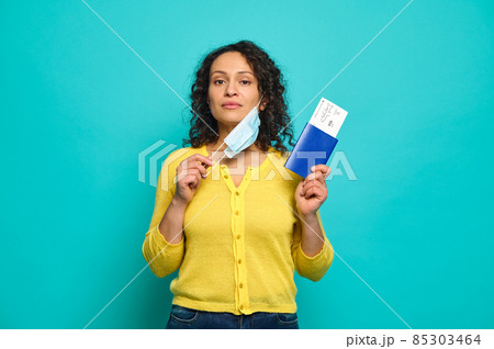 Beautiful mid adult woman traveler in yellow sweater puts her medical mask off and confidently looks at camera, holding a boarding pass, air flight ticket and passport. Copy ad space. Travel concept Beautiful mid adult woman traveler in yellow sweater puts her medical mask off and confidently looks at camera, holding a boarding pass, air flight ticket and passport. Copy ad space. Travel concept 85303464