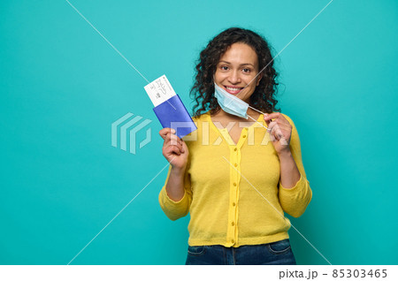 Charming woman traveler in yellow sweater putting her medical mask off and smiles with cheerful toothy smile looking at camera, holding a boarding pass, air flight ticket and passport. Copy space Charming woman traveler in yellow sweater putting her medical mask off and smiles with cheerful toothy smile looking at camera, holding a boarding pass, air flight ticket and passport. Copy space 85303465