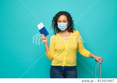 Cheerful Caucasian woman with curly dark hair, wearing protective medical mask, poses with a boarding pass and air ticket in the passport and a suitcase. Concept of safety travel during pandemic 85303467
