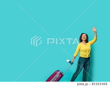 Full length of beautiful woman in casual clothes, waving hand, holding identity document and boarding pass, posing with suitcase, smiling looking to the side at a blue background with copy space 85303468