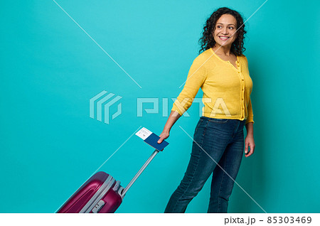 Happy curly dark-haired woman, traveler in bright yellow sweater and blue jeans holding passport with boarding pass and air ticket, smiles, posing with luggage on blue background with copy space 85303469