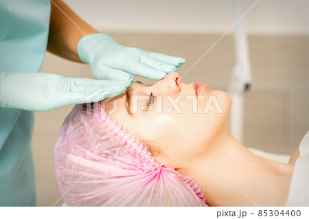 Face peeling at the beautician. Facial treatment. The beautician applies a cleansing face mask to the female patient. Face peeling at the beautician. Facial treatment. The beautician applies a cleansing face mask to the female patient. 85304400