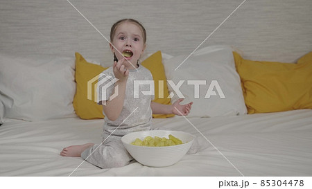 little girl eating golden potato chips sitting on the bed in the bedroom, fast food for kid, unhealthy baby food, enjoying the taste of potato spices, girl's favorite snack at home, happy daughter little girl eating golden potato chips sitting on the bed in the bedroom, fast food for kid, unhealthy baby food, enjoying the taste of potato spices, girl's favorite snack at home, happy daughter 85304478