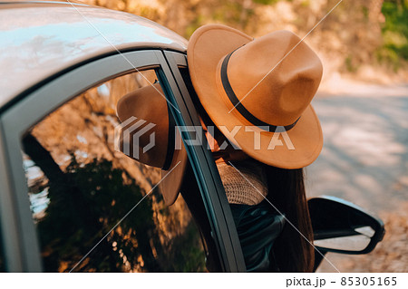 Beautiful young woman carrying hat and looking happy while enjoying road trip. Young woman with long hair in brown hat and sweater looking out from white car holding hat and scarf against autumn 85305165