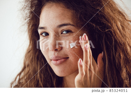 Portrait of woman with curly hair and freckles applying moisturizer to her face. 85324051
