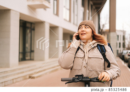 Portrait of female courier food delivery with big thermo backpack on back talking on smartphone with client standing with electric scooter in urban street. Portrait of female courier food delivery with big thermo backpack on back talking on smartphone with client standing with electric scooter in urban street. 85325486