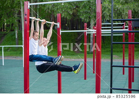 Caucasian man trains a boy on the horizontal bar on the playground. Dad and son go in for outdoor sports. Caucasian man trains a boy on the horizontal bar on the playground. Dad and son go in for outdoor sports. 85325596