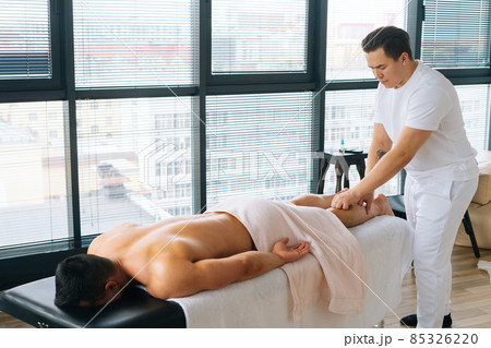 Professional male masseur with strong tattooed hands massaging lower leg of muscular athlete man after physical sports workout 85326220