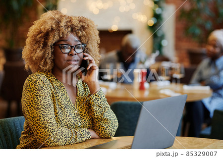 African woman in eyeglasses talking on mobile phone while sitting at the table with laptop in coffee shop 85329007