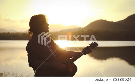 Man playing acoustic guitar outdoors with sunlight reflected on water surface at sunset lake. Man playing acoustic guitar outdoors with sunlight reflected on water surface at sunset lake. 85339024