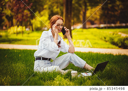 Redhead young woman works with laptop and cellphone, outdoors while sitting on the grass. Freelance  85345498