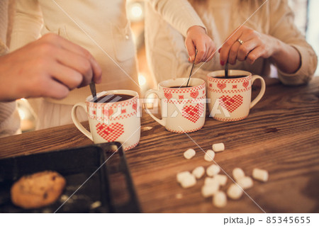 Family: mom, dad and daughter in white sweaters cook and drink cocoa with marshmallows. Closeup hands and cups. Christmas concept. 85345655