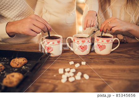 Family: mom, dad and daughter in white sweaters cook and drink cocoa with marshmallows. Closeup hands and cups. Christmas concept. 85345656