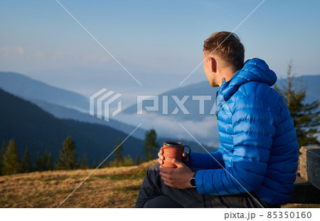 Hiking in the mountains. Guy sitting outdoors on mountain meadow at sunny morning, looking at very beautiful misty mountain hills. Active trekking recreation. 85350160
