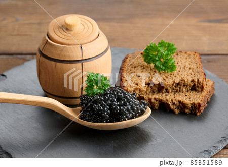 fresh grainy black paddlefish caviar in brown wooden spoon on a brown table, close up fresh grainy black paddlefish caviar in brown wooden spoon on a brown table, close up 85351589