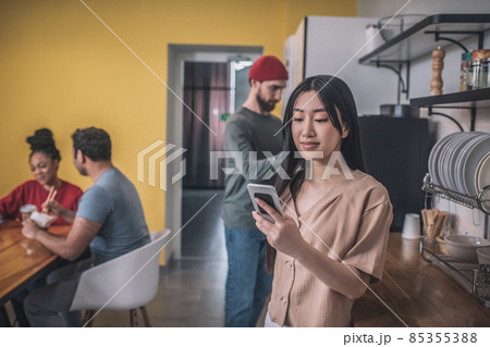 Asian girl with smartphone and colleagues behind in kitchen 85355388