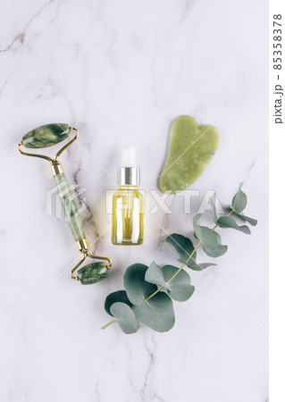 Vertical flat lay composition with jade gua sha facial roller with beauty serum oil on white marble table background with copy space. Facial massage kit for lifting massage therapy from natural stones 85358378