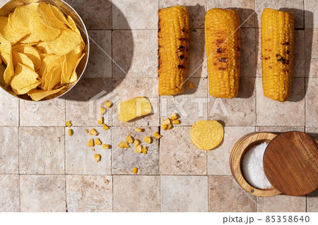 Delicious organic homemade grilled corn cobs and bowl of nacho chips with salt cellar on earth toned stone tales background top view 85358640