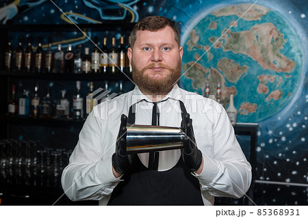 A bearded adult man of European appearance, a professional bartender, holds in his hands in black latex gloves a tool for preparing and mixing alcoholic cocktails in a nightclub 85368931