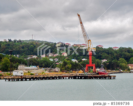 A tall floating crane stands on the pier in the sea near the ship against the backdrop of green hills and houses 85369736