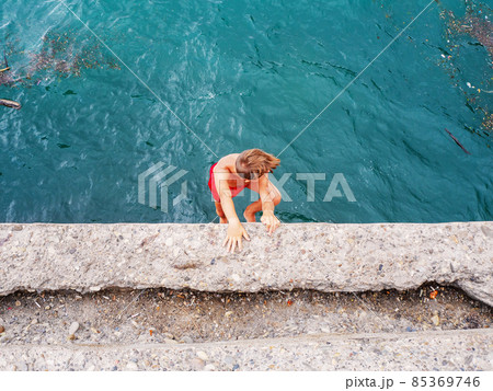 A boy in red swimming trunks descends from a concrete pier into the sea water A boy in red swimming trunks descends from a concrete pier into the sea water 85369746