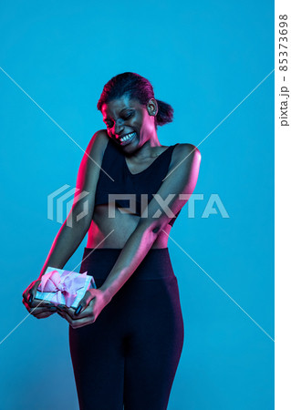 Happy young beautiful dark skinned woman with gift box isolated on blue studio background in neon light. Happy young beautiful dark skinned woman with gift box isolated on blue studio background in neon light. 85373698