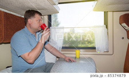 A full grown man in a train compartment is sitting at a table with a mug of tea, the concept of lunch on a journey 85374879