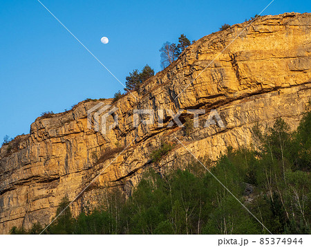 White moon is above the rock. Rock against the evening blue sky. High peaked sharp rock. Rocky sharp mountain ridge with a chasm. 85374944
