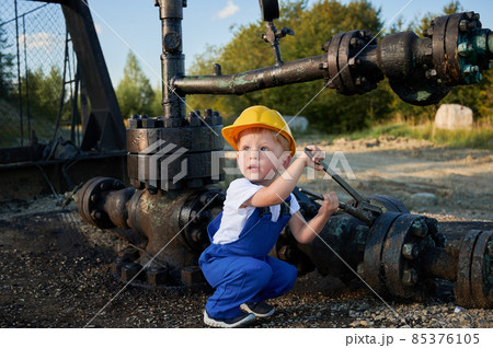 Little boy in yellow helmet studying mechanics. Child sat down, trying to turn nut on pipeline with wrench outdoors. Creative acquaintance of child with working tools. 85376105
