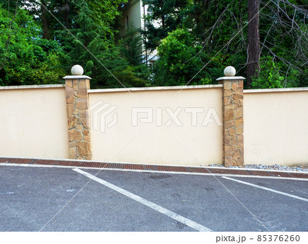 A fence running along a marked asphalt road section and green trees and a fragment of a house behind the fence A fence running along a marked asphalt road section and green trees and a fragment of a house behind the fence 85376260