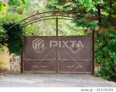 A brown iron gate with a wrought golden pattern and a patterned arch above it and thickets of vegetation next to and behind them A brown iron gate with a wrought golden pattern and a patterned arch above it and thickets of vegetation next to and behind them 85376269