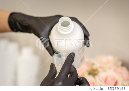 A hairdresser in black gloves is preparing hair dye with a bottle in a hair salon, close up. 85379143