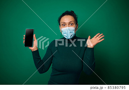 Cheerful happy millennial woman in protective face cover looks at camera and shows her smartphone with empty blank screen for copy space, isolated over green background Cheerful happy millennial woman in protective face cover looks at camera and shows her smartphone with empty blank screen for copy space, isolated over green background 85380795